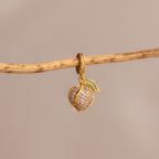 The Alexandrite Peach Charm features a gold peach-shaped pendant with pavé stones, suspended from a branch on a beige background—a delightful addition to any jewelry collection.