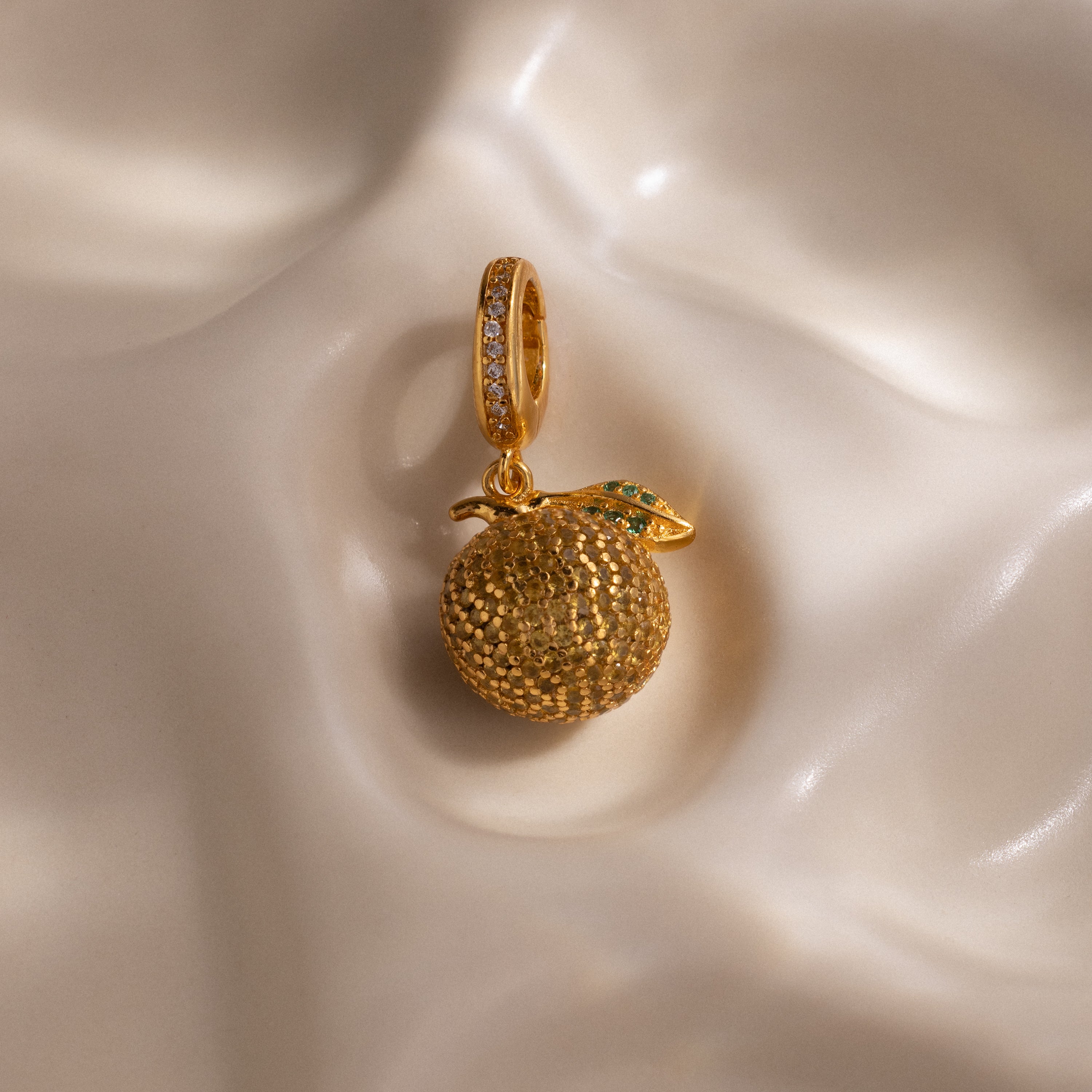 The Topaz Citrus Charm features a gold-textured orange pendant with pavé accents and a jeweled leaf, set against a soft, wavy cream background—a stunning addition to any jewelry collection.