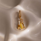 The Diamond Banana Charm features a gold, banana-shaped design with pavé stones, shown on a smooth cream surface—a playful and unique addition to any jewelry collection.