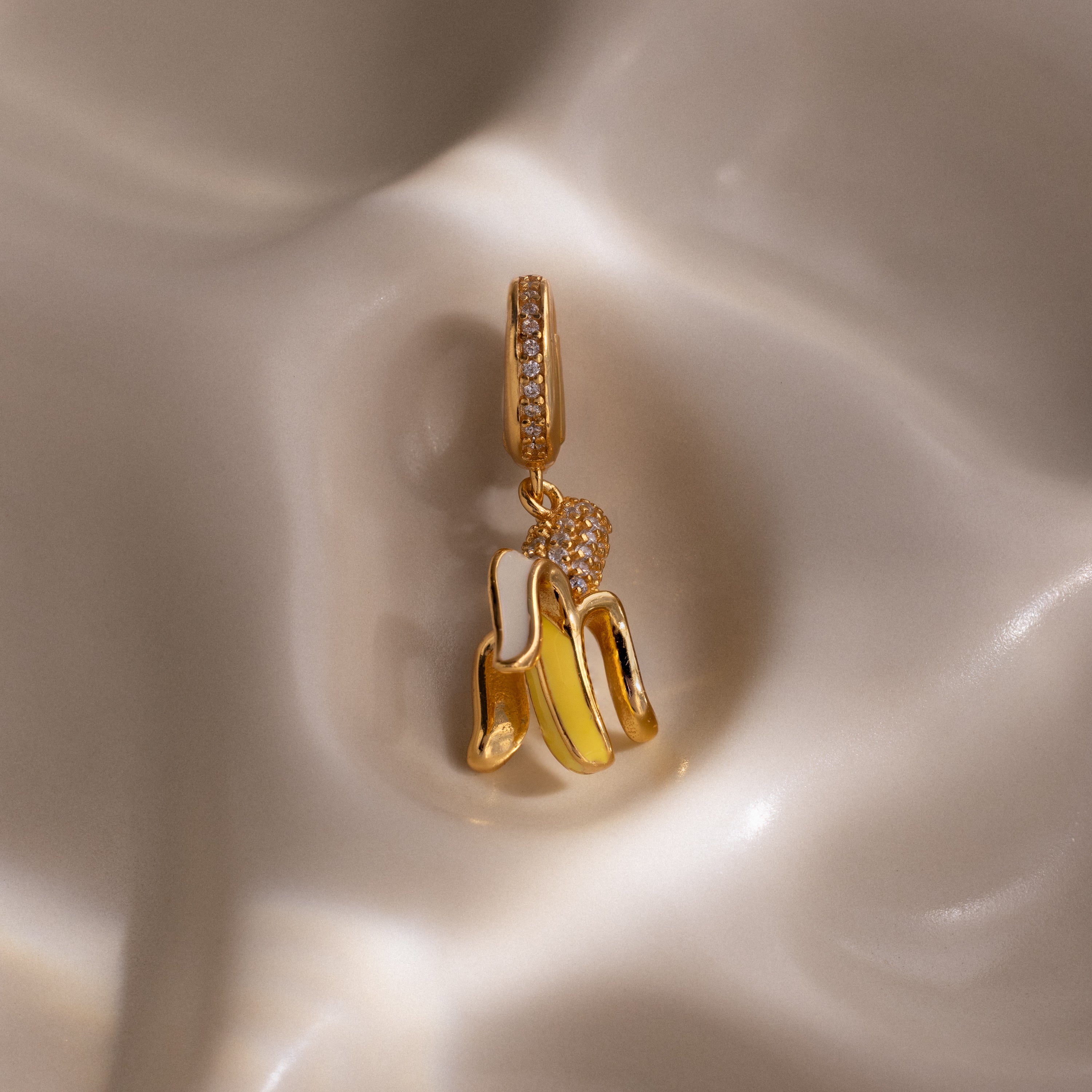 The Diamond Banana Charm features a gold, banana-shaped design with pavé stones, shown on a smooth cream surface—a playful and unique addition to any jewelry collection.