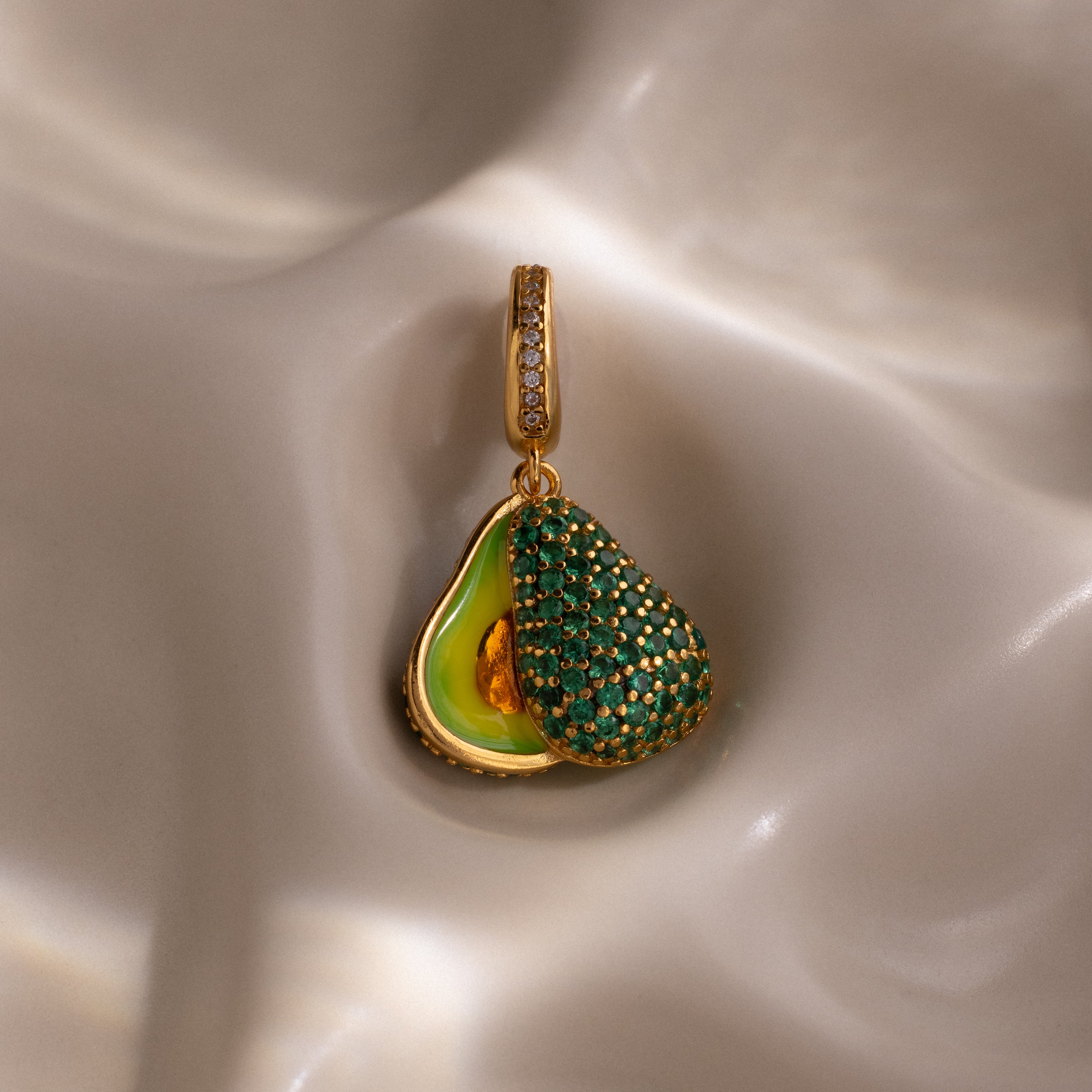 The Emerald Avocado Charm features pavé stones and green gemstones in a gold avocado-shaped pendant, displayed on silky cream-colored fabric—a unique addition to any jewelry collection.