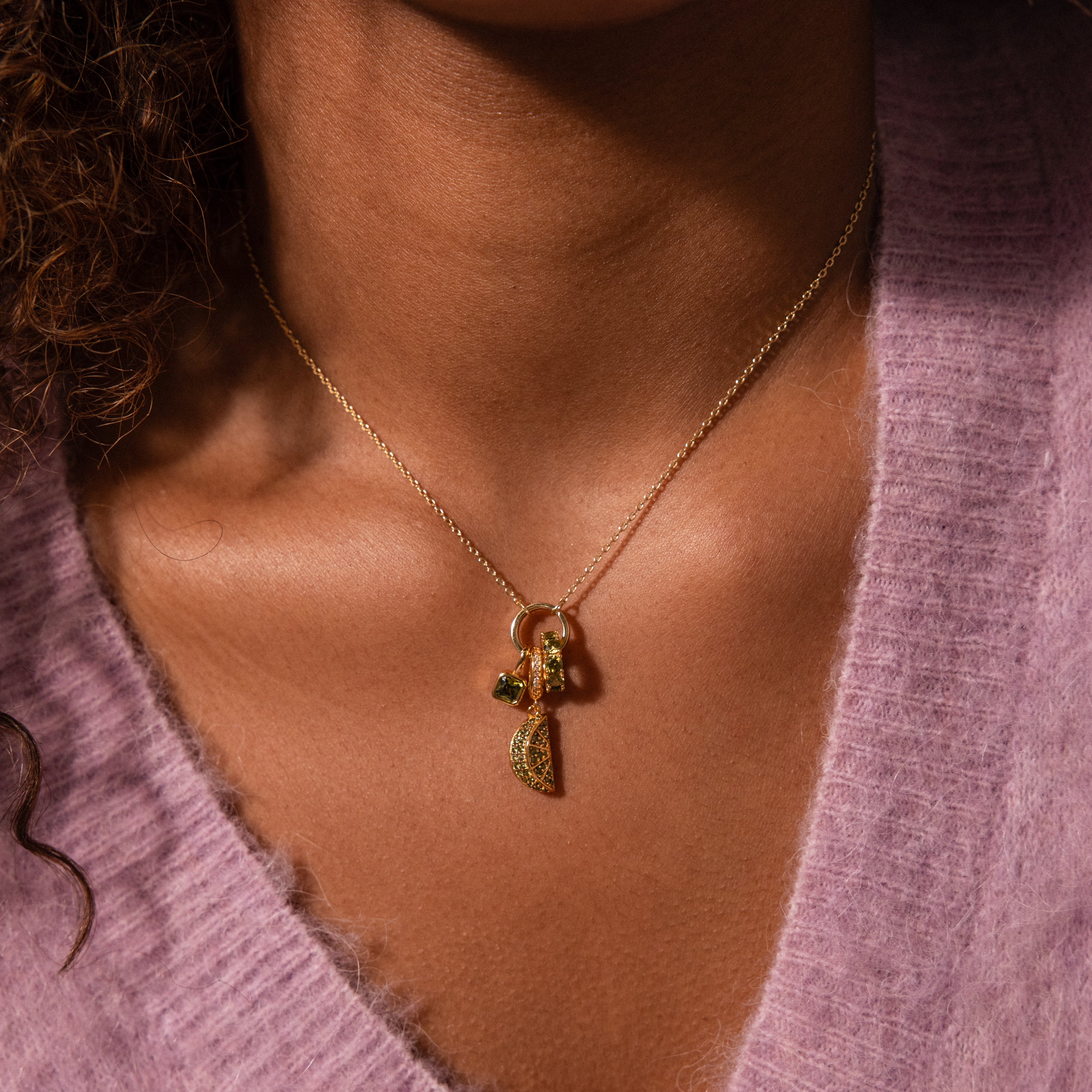 A close-up of a person wearing a Sapphire Blackberry Charm necklace adorned with pavé stones and a soft purple V-neck sweater from their jewelry collection.