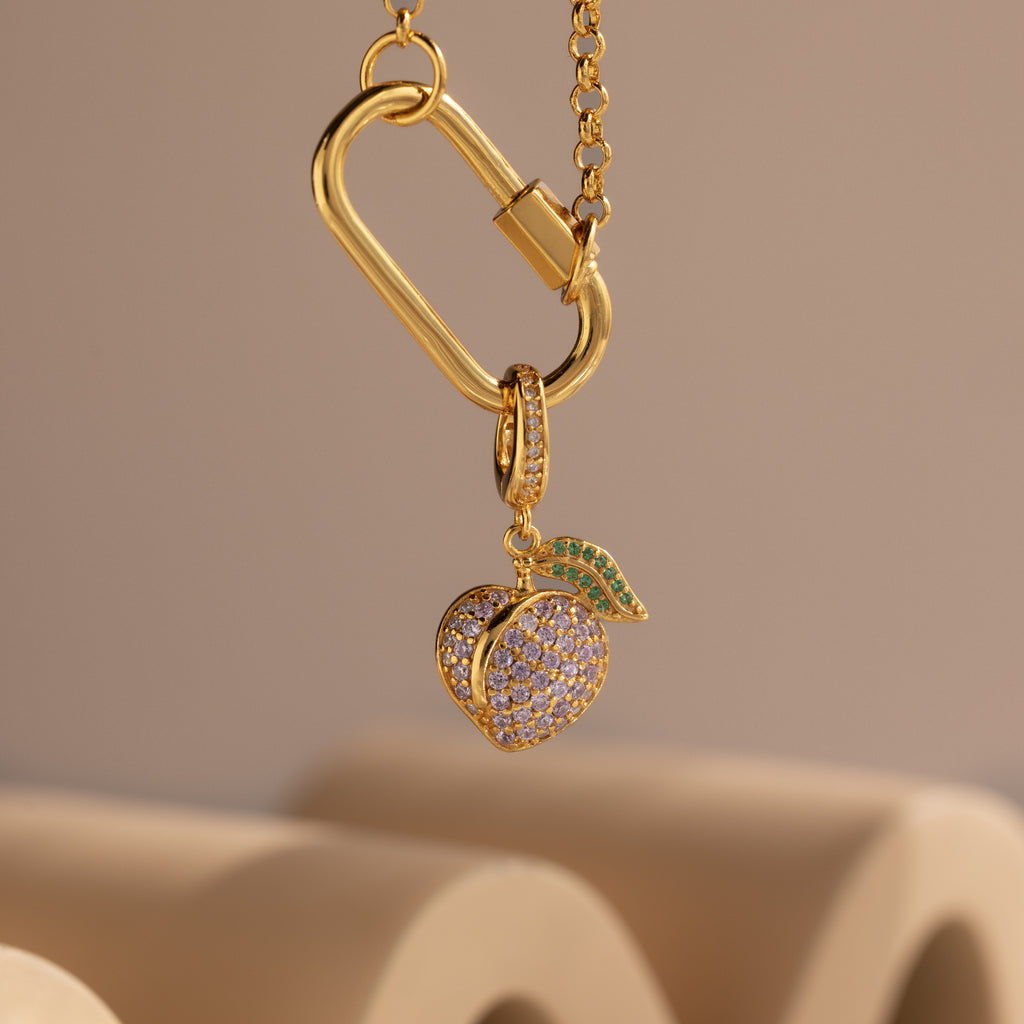Alexandrite Peach Charm gold necklace features a sparkling peach-shaped pendant with pavé stones and a green jeweled leaf, showcased against a beige background.