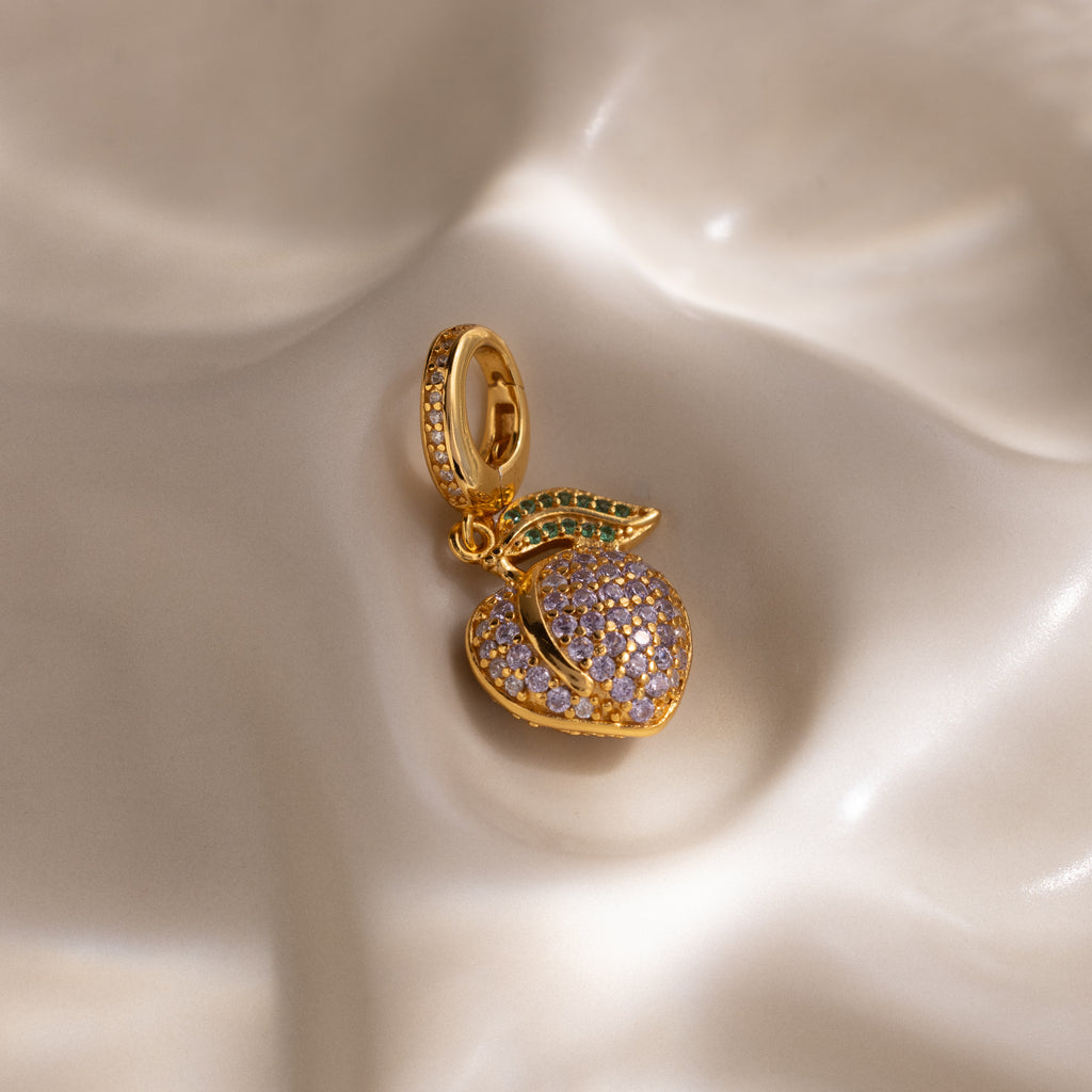 The Alexandrite Peach Charm features a gold peach shape with pavé purple and green stones on a smooth, creamy base—a delightful addition to any jewelry collection.