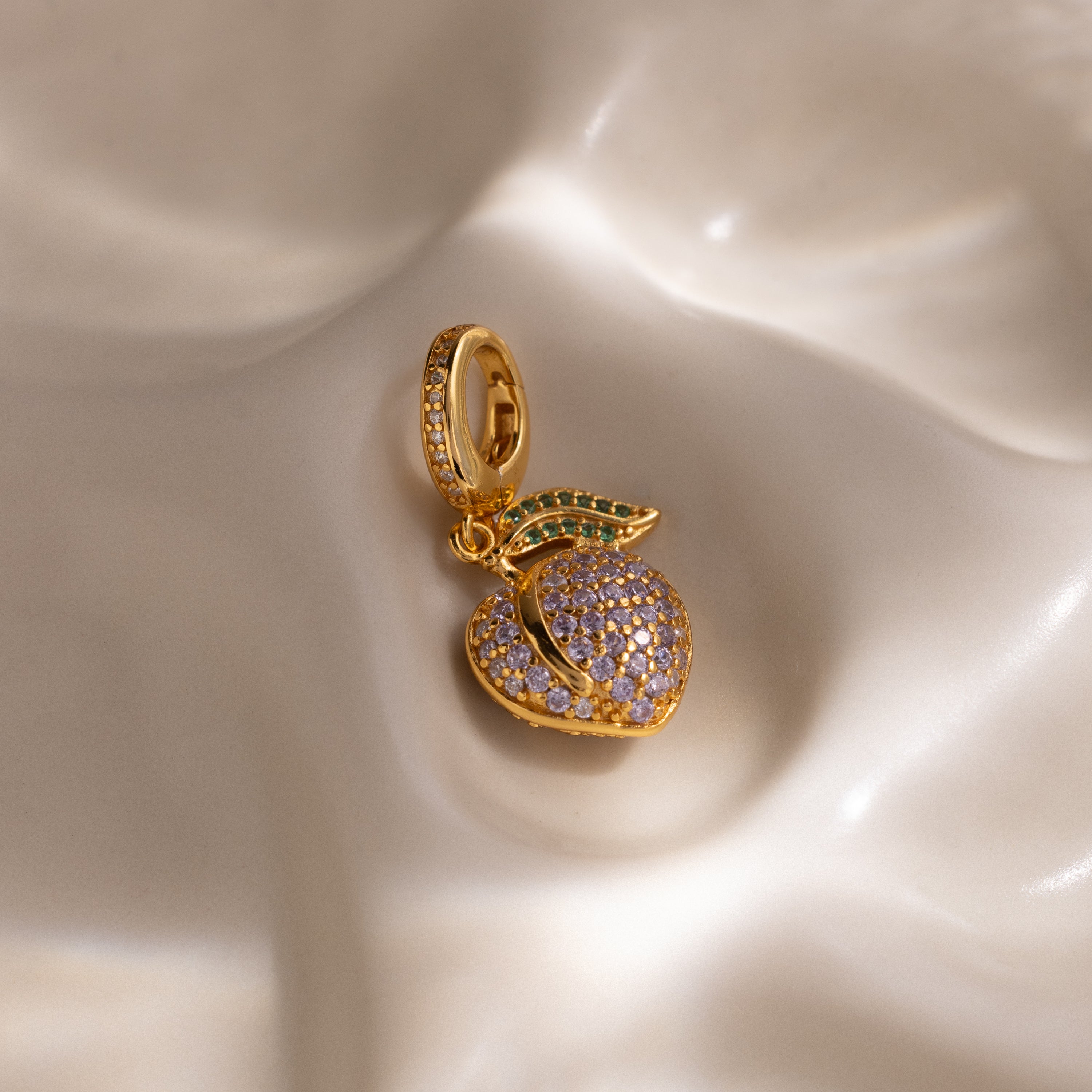 The Alexandrite Peach Charm features a gold peach shape with pavé purple and green stones on a smooth, creamy base—a delightful addition to any jewelry collection.