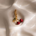 The Ruby Cherry Charm features red enamel cherries, pavé stones, and a green leaf detail, shown on silky cream fabric—a standout piece from our jewelry collection.