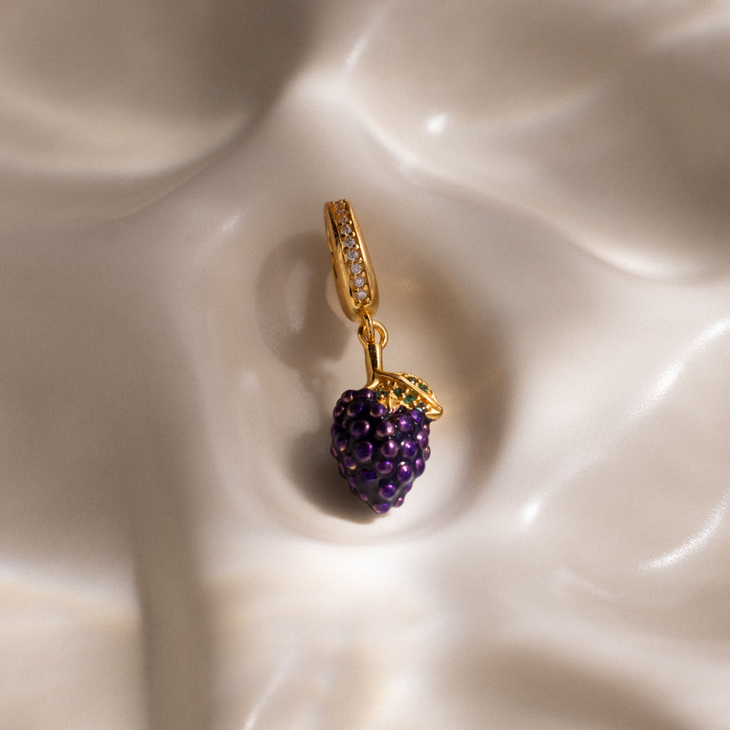 The Amethyst Grape Charm, shaped like a cluster of gold and purple grapes, rests on a smooth cream-colored surface, adding playful elegance to any jewelry collection.