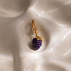 The Amethyst Grape Charm, shaped like a cluster of gold and purple grapes, rests on a smooth cream-colored surface, adding playful elegance to any jewelry collection.
