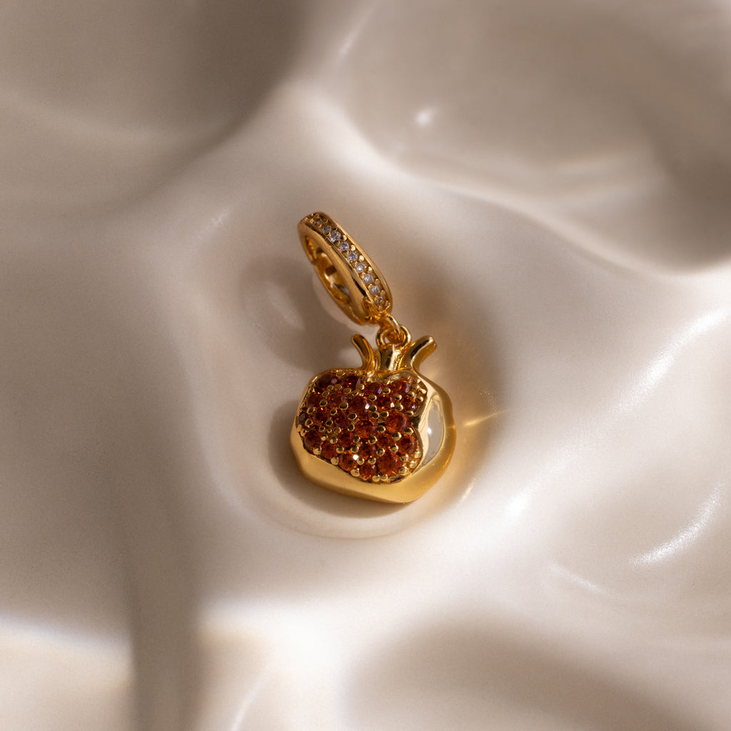 The Garnet Pomegranate Charm features red pavé stones set in a gold pendant, elegantly displayed on cream-colored fabric—a charming addition to any jewelry collection.