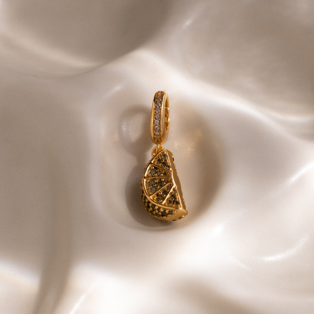 The Peridot Lime Charm, shaped like a lemon slice with pavé stones, sparkles atop creamy fabric—a standout piece from our jewelry collection.
