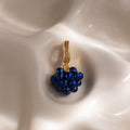 The Sapphire Blackberry Charm, a gold pendant adorned with blue beads and small crystals, is featured from our jewelry collection against a softly textured cream background.