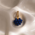 The Sapphire Blackberry Charm, a gold pendant adorned with blue beads and small crystals, is featured from our jewelry collection against a softly textured cream background.