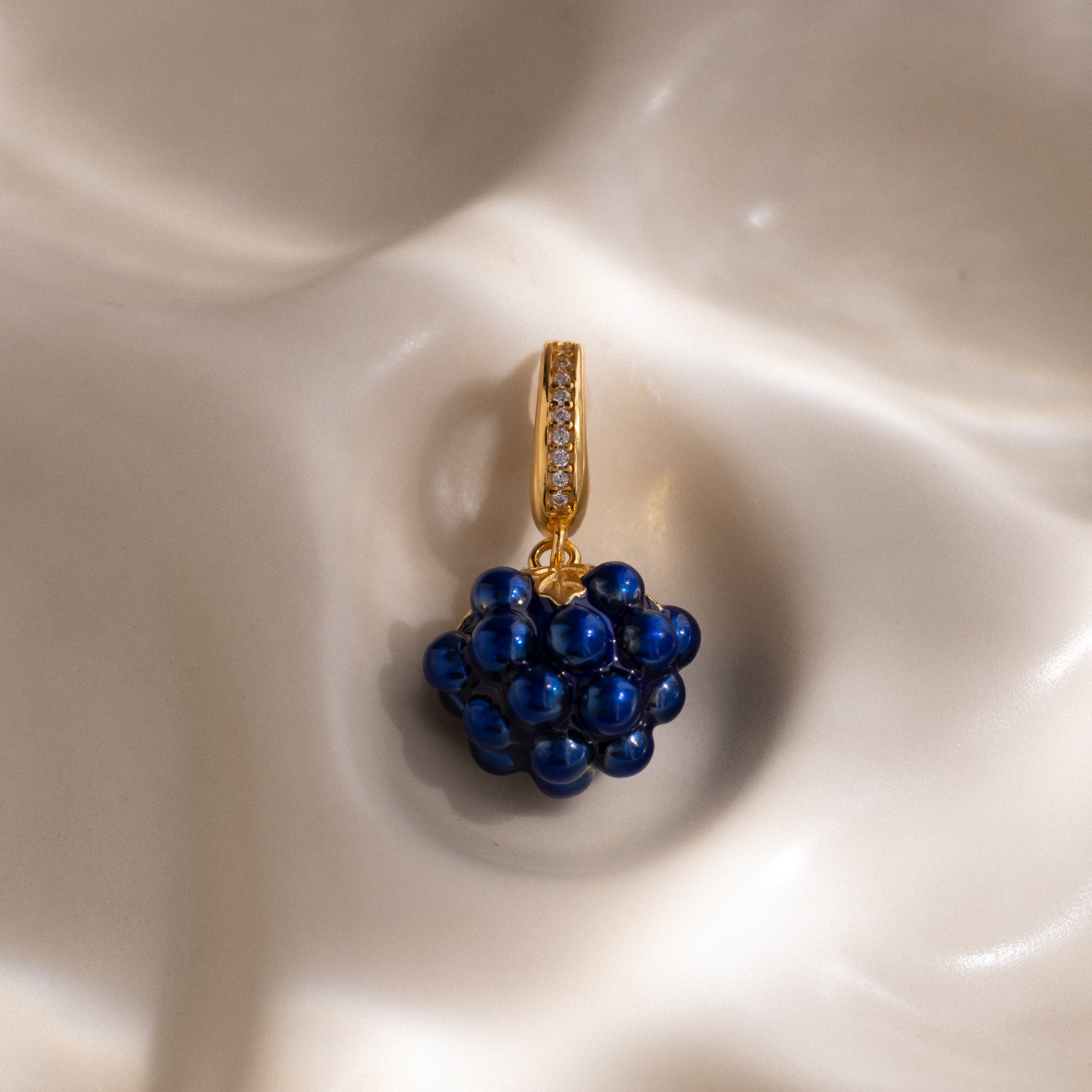The Sapphire Blackberry Charm, a gold pendant adorned with blue beads and small crystals, is featured from our jewelry collection against a softly textured cream background.