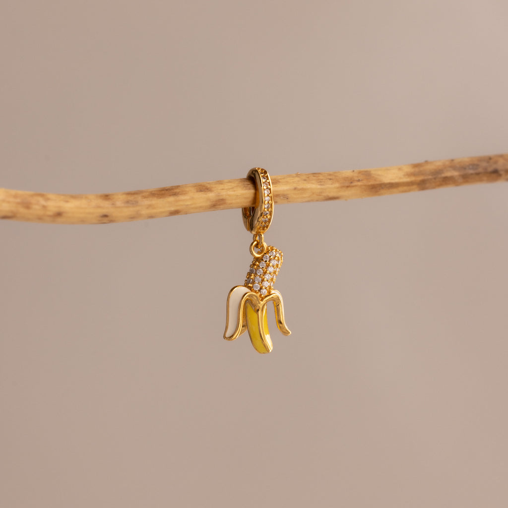 The Diamond Banana Charm, featuring pavé stones in a gold banana shape, hangs elegantly from a thin branch on a beige background—an eye-catching addition to any jewelry collection.