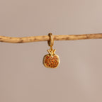 The Garnet Pomegranate Charm, featuring red pavé stones, hangs from a branch against a neutral background.