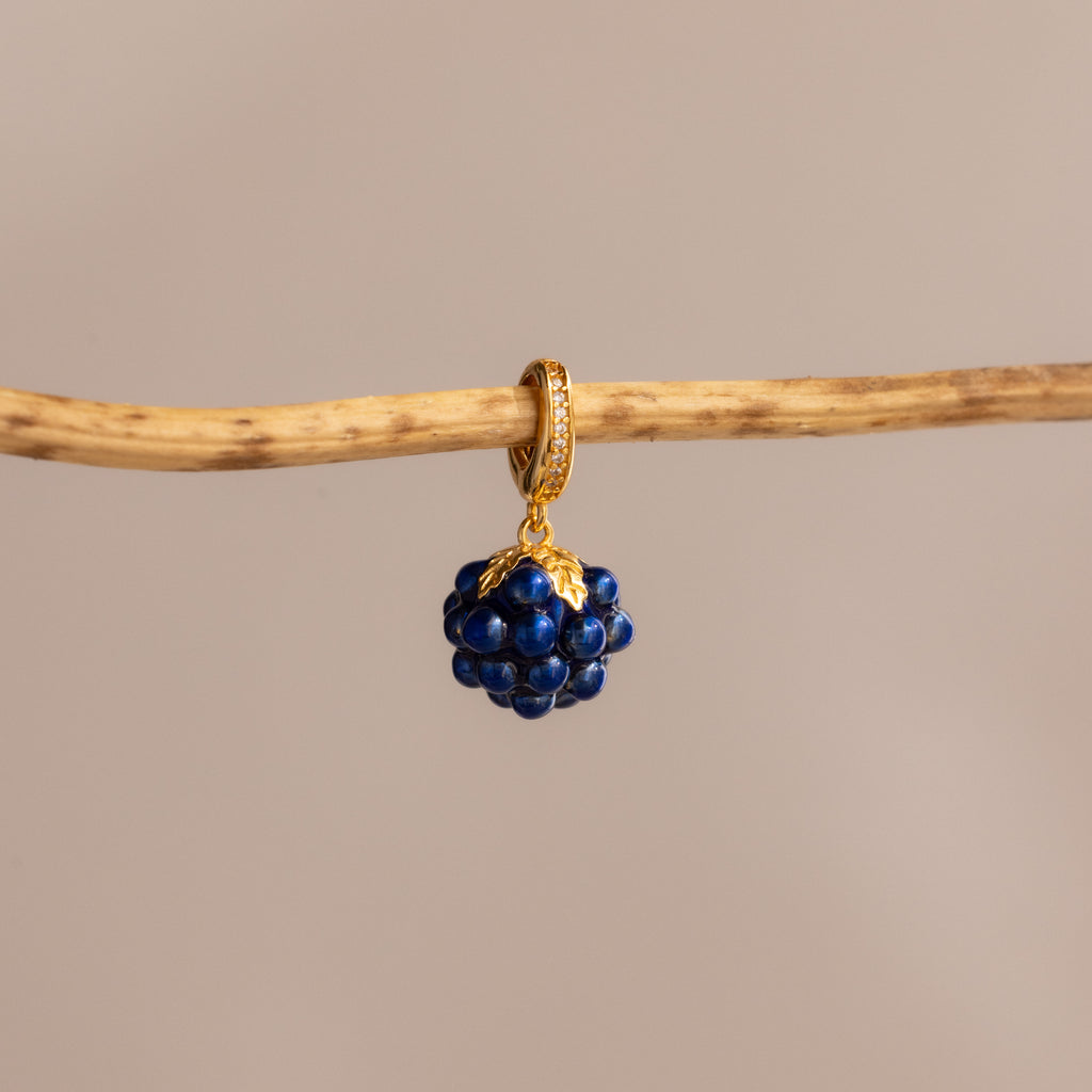 The Sapphire Blackberry Charm, a gold hoop earring adorned with dark blue beads, hangs elegantly on a thin wooden branch against a beige background—an eye-catching piece from our jewelry collection.