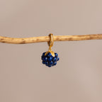 The Sapphire Blackberry Charm, a gold hoop earring adorned with dark blue beads, hangs elegantly on a thin wooden branch against a beige background—an eye-catching piece from our jewelry collection.