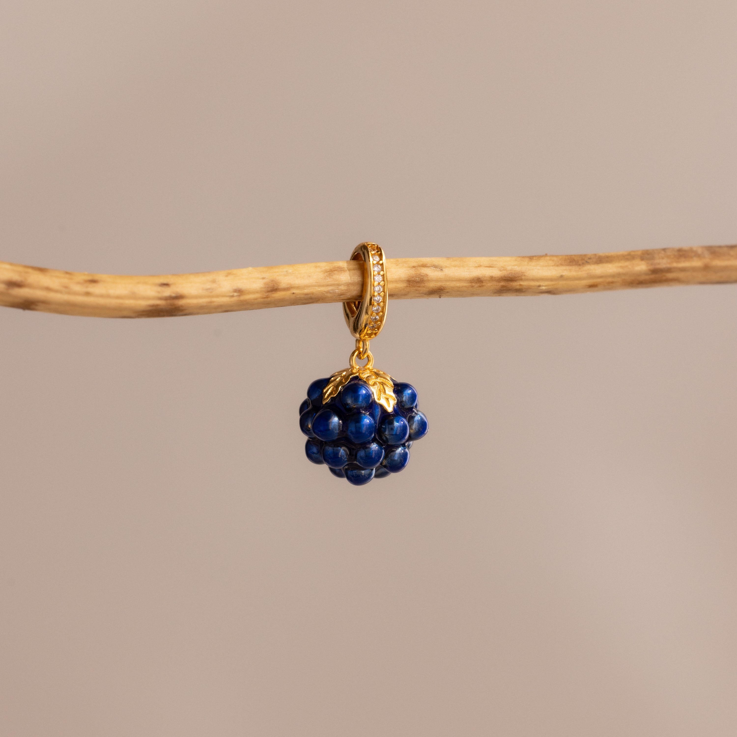 The Sapphire Blackberry Charm, a gold hoop earring adorned with dark blue beads, hangs elegantly on a thin wooden branch against a beige background—an eye-catching piece from our jewelry collection.