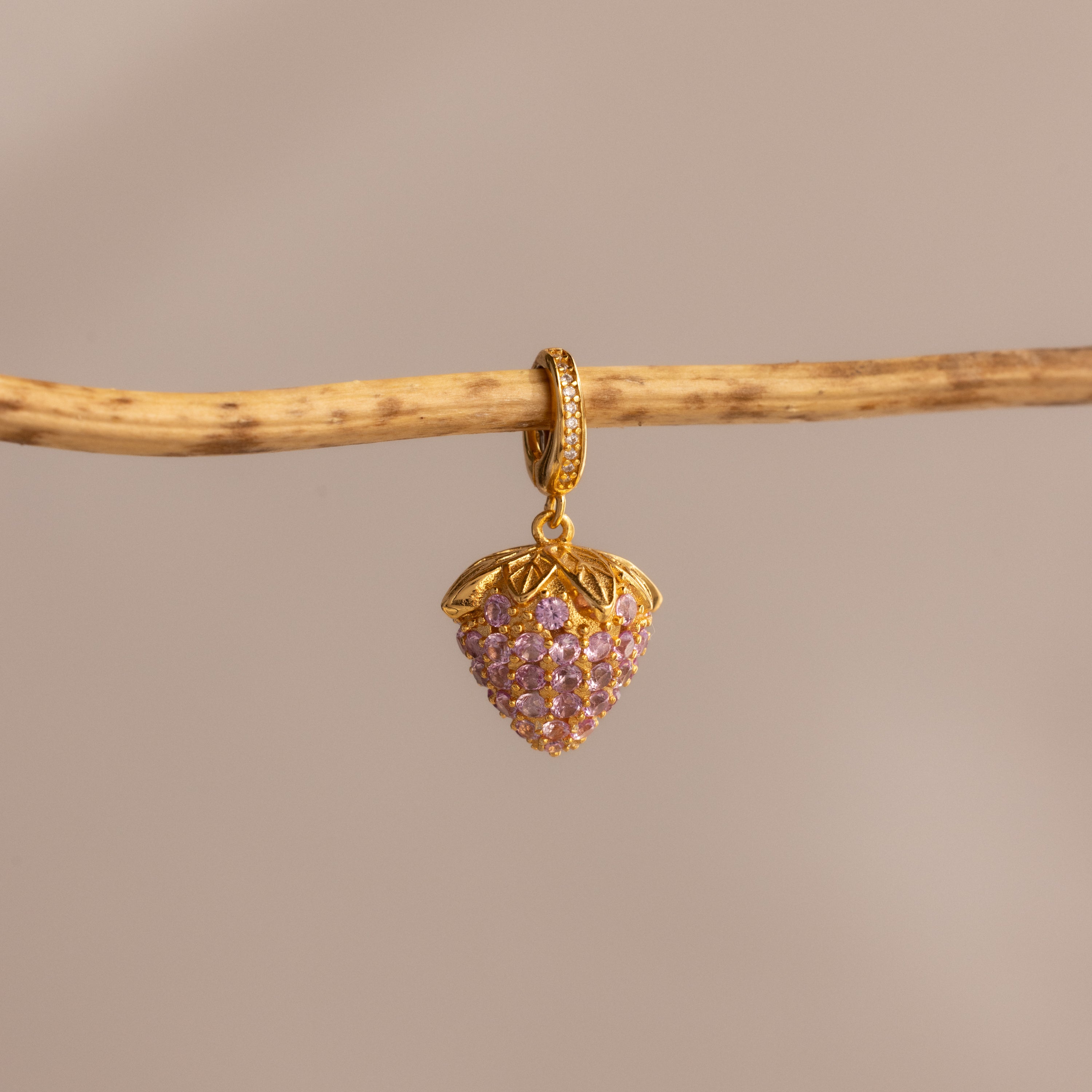 The Pink Tourmaline Raspberry Charm, featuring a gold raspberry-shaped pendant with pavé stones and pink tourmaline gems, hangs from a twig against a beige background—a unique piece from our jewelry collection.