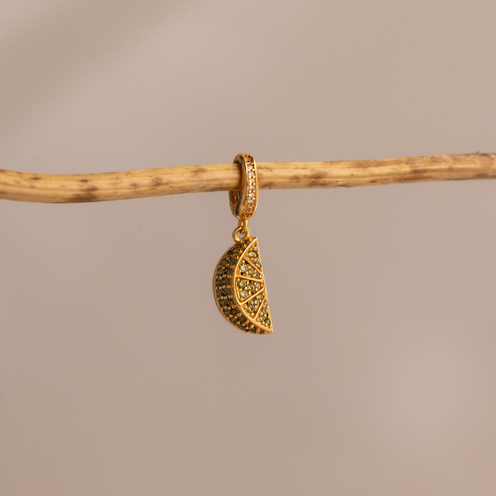 The Peridot Lime Charm features sparkling pavé stones and hangs from a branch against a beige background—a vibrant, eye-catching addition to any jewelry collection.