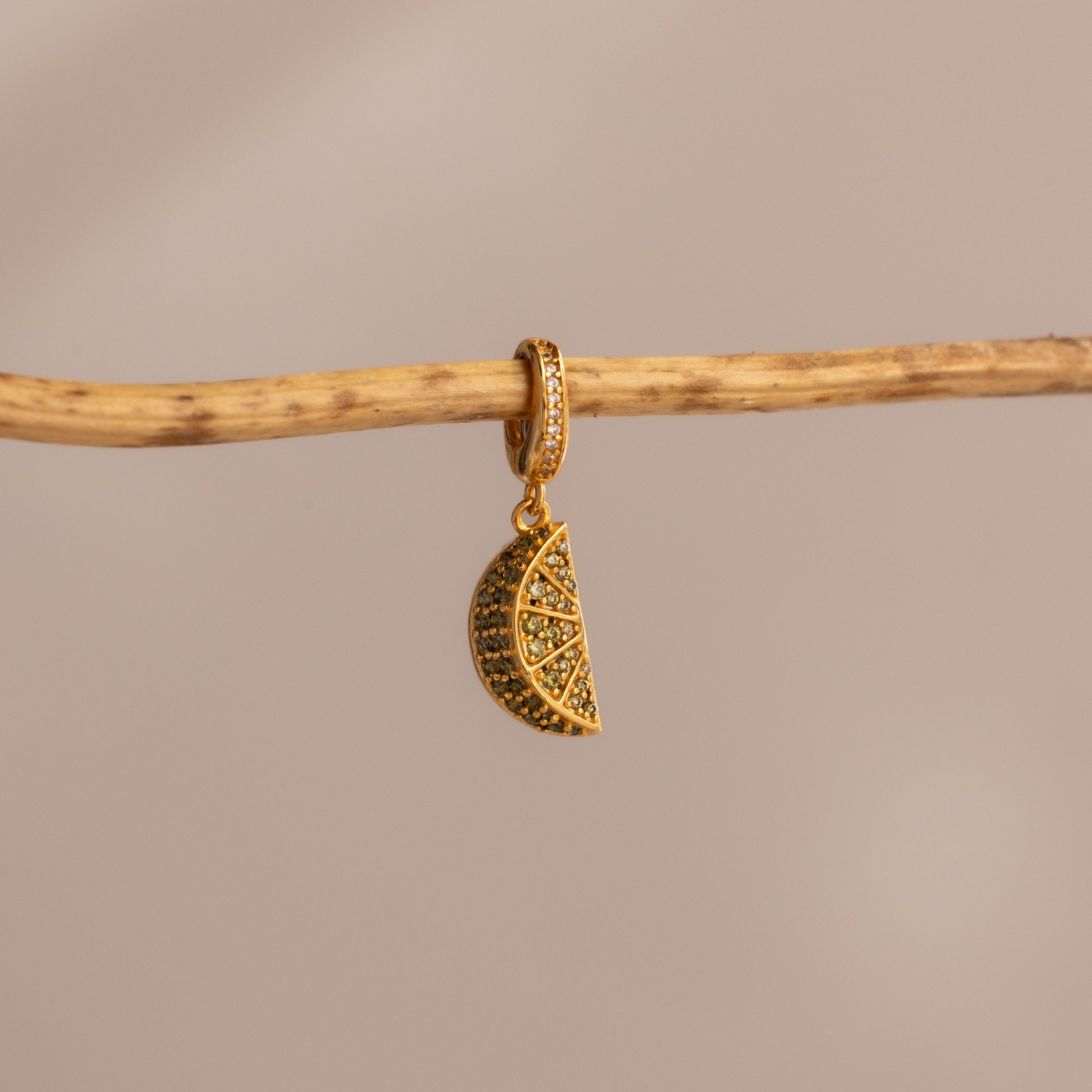 The Peridot Lime Charm features sparkling pavé stones and hangs from a branch against a beige background—a vibrant, eye-catching addition to any jewelry collection.