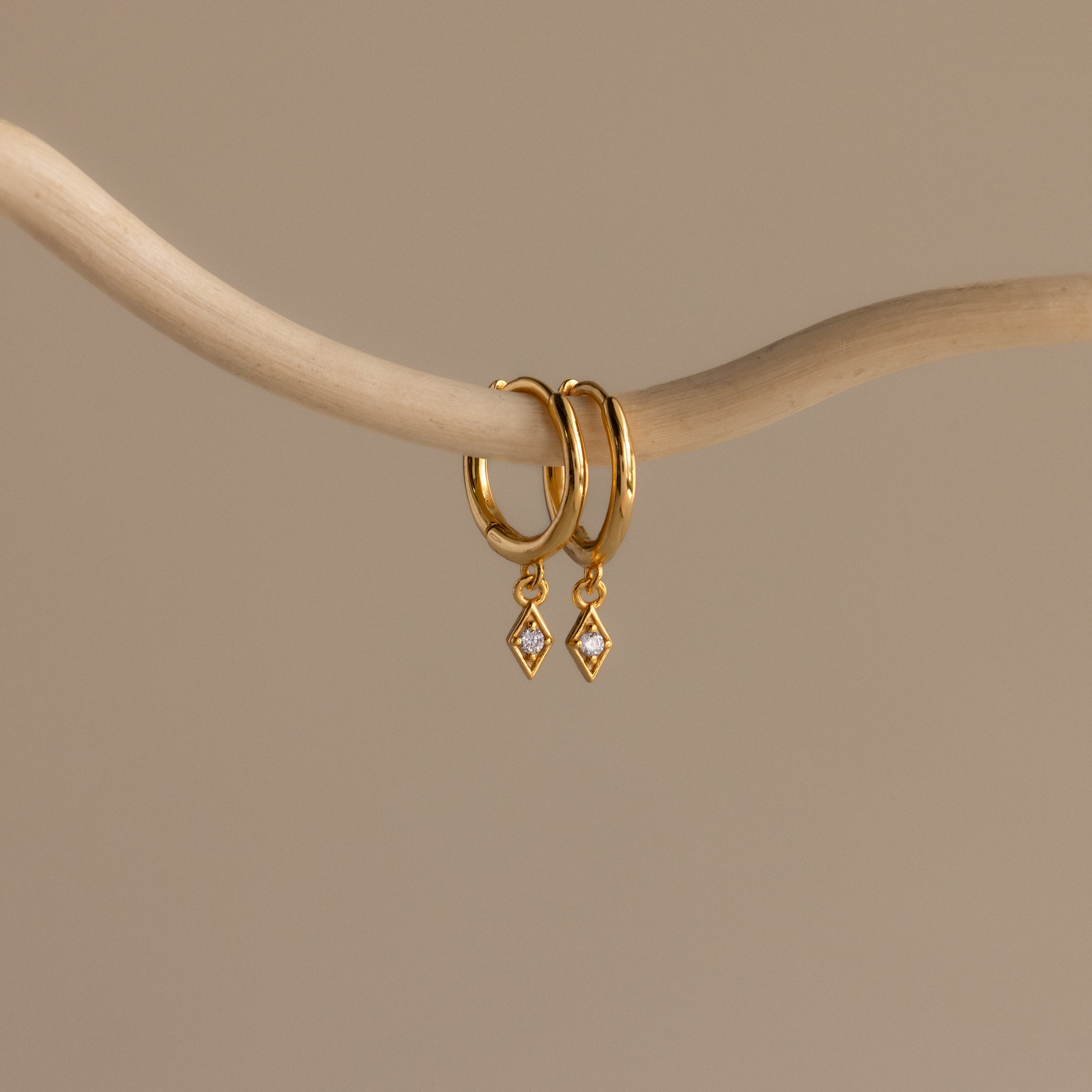 The Ericka Birthstone Huggies in Diamond / April hang from a curved beige branch on a neutral background—an elegant choice for anyone who loves diamond-accented or personalized earrings.