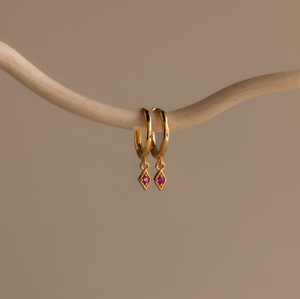 The Ericka Ruby Huggies are gold hoop earrings featuring small red diamond-shaped pendants, elegantly displayed on a curved beige branch against a neutral background.