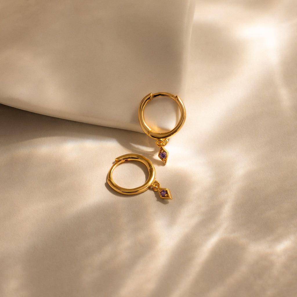 Ericka Birthstone Huggies in Amethyst (February): two gold hoop earrings with small purple gemstone charms, beautifully displayed on soft, sunlit beige fabric.