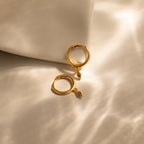 Ericka Birthstone Huggies in Amethyst (February): two gold hoop earrings with small purple gemstone charms, beautifully displayed on soft, sunlit beige fabric.