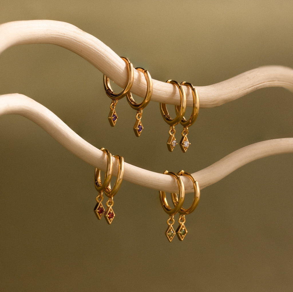 Ericka Opal Huggies feature gold hoops with opal gemstone charms, hanging elegantly on light curved branches. Personalize your style with these timeless earrings that add a subtle shimmer to any look.