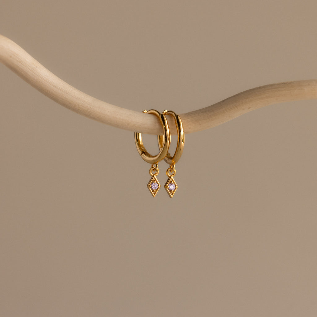 Ericka Alexandrite Huggies evoke the allure of diamond charm earrings, elegantly hanging from a smooth, curved beige branch against a plain background.