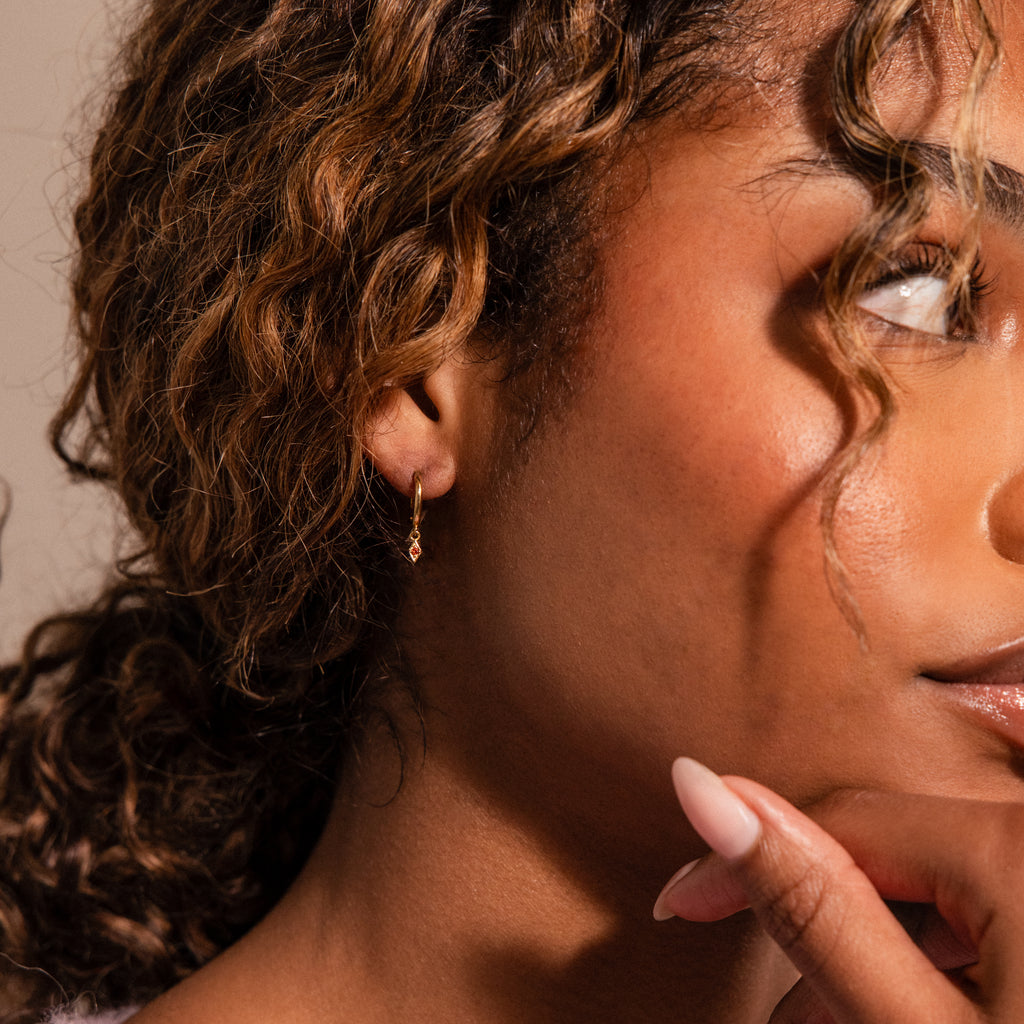 A woman with curly hair touches her chin, showcasing the Ericka Sapphire Huggies—small gold personalized jewelry earrings—in her ear.