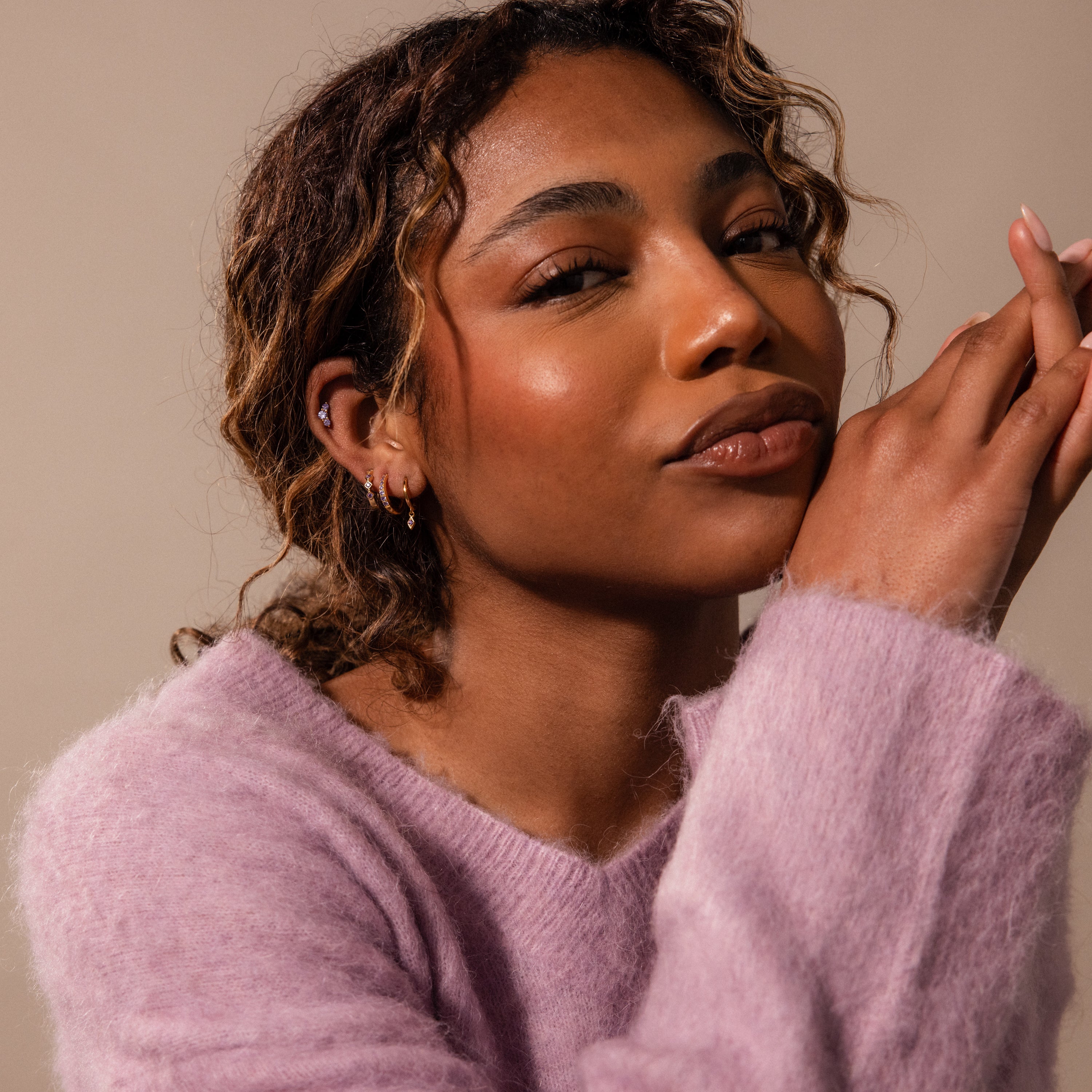 A woman with curly hair in a soft pink sweater confidently showcases Ericka Amethyst Huggies, her hands near her face drawing attention to the elegant birthstone earrings.