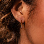 Close-up of an ear wearing Ericka Topaz Huggies, small gold hoops with a dangling purple gemstone charm—ideal for fans of personalized jewelry.