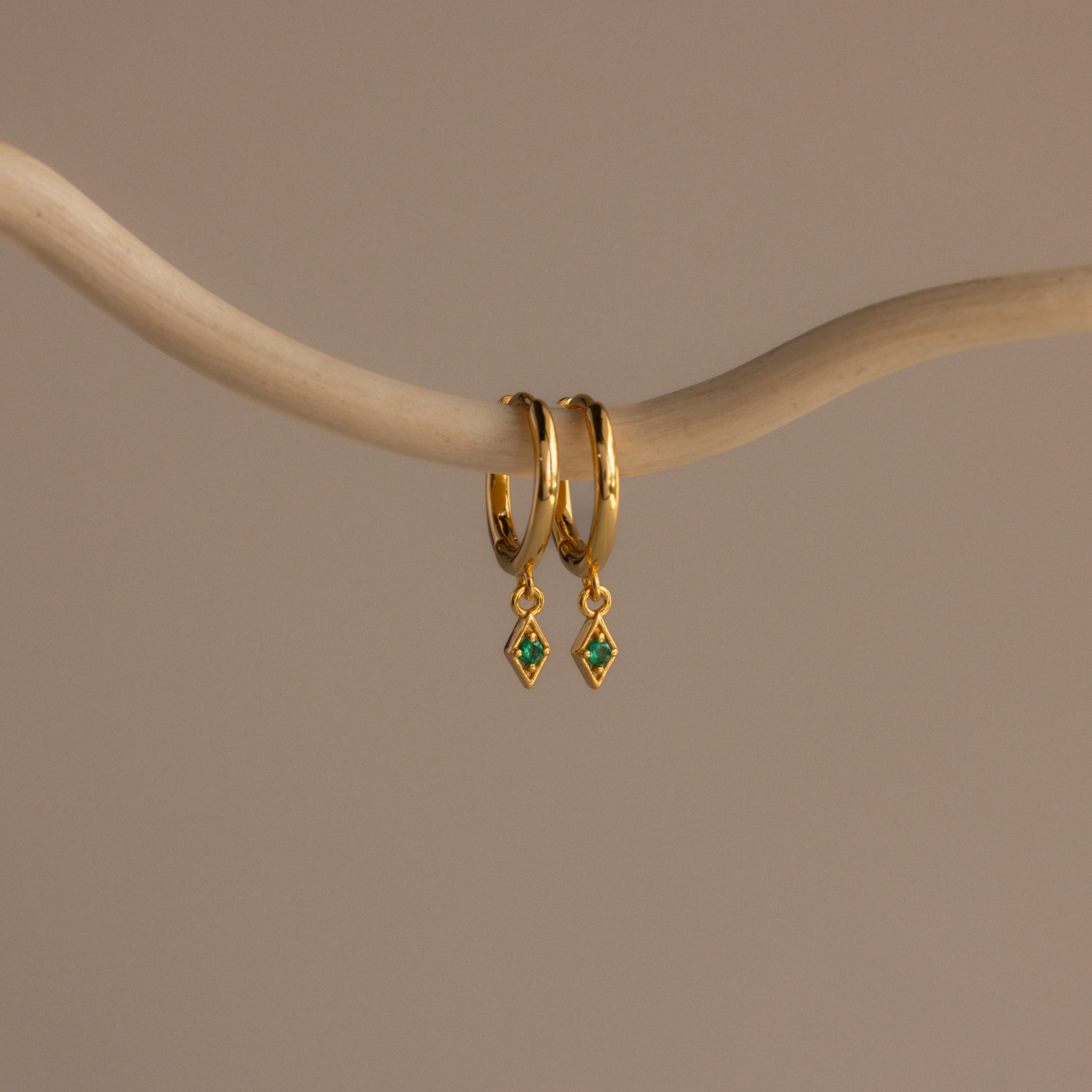 A pair of Ericka Birthstone Huggies in emerald (May), featuring gold hoops with small green diamond-shaped charms, hang from a curved beige branch.