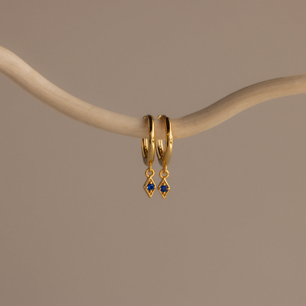 The Ericka Sapphire Huggies are gold hoop earrings featuring delicate blue diamond-shaped charms, elegantly displayed on a curved beige branch—perfect birthstone jewelry to personalize any look.