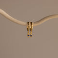 The Ericka Sapphire Huggies are gold hoop earrings featuring delicate blue diamond-shaped charms, elegantly displayed on a curved beige branch—perfect birthstone jewelry to personalize any look.