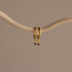The Ericka Sapphire Huggies are gold hoop earrings featuring delicate blue diamond-shaped charms, elegantly displayed on a curved beige branch—perfect birthstone jewelry to personalize any look.