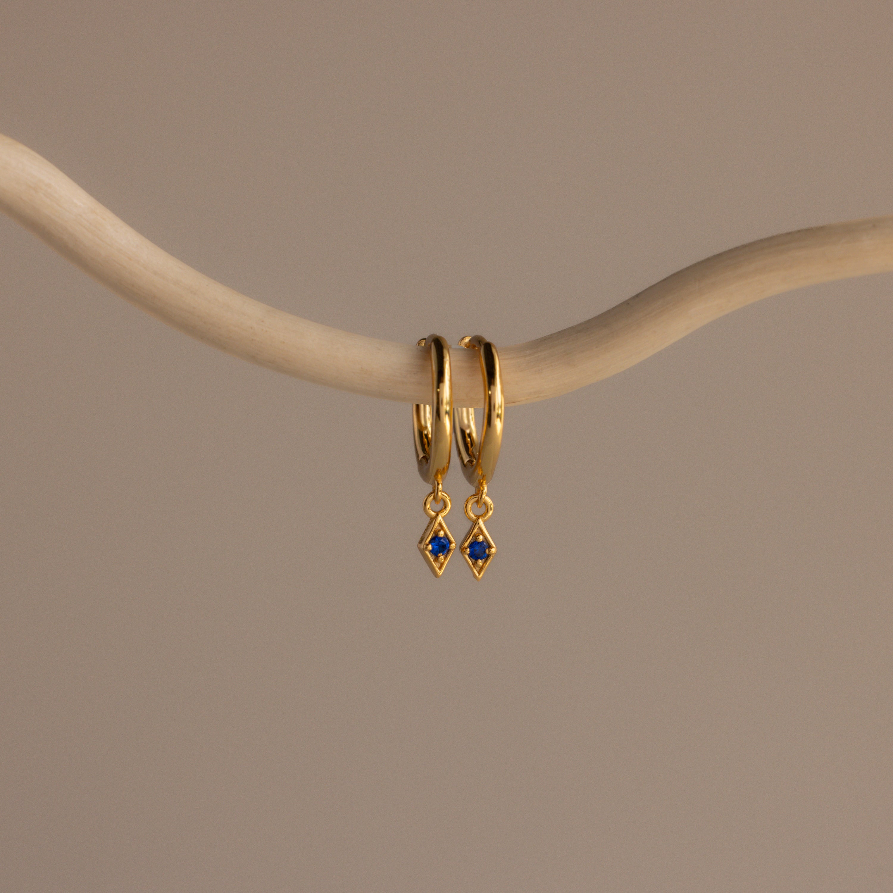 The Ericka Sapphire Huggies are gold hoop earrings featuring delicate blue diamond-shaped charms, elegantly displayed on a curved beige branch—perfect birthstone jewelry to personalize any look.
