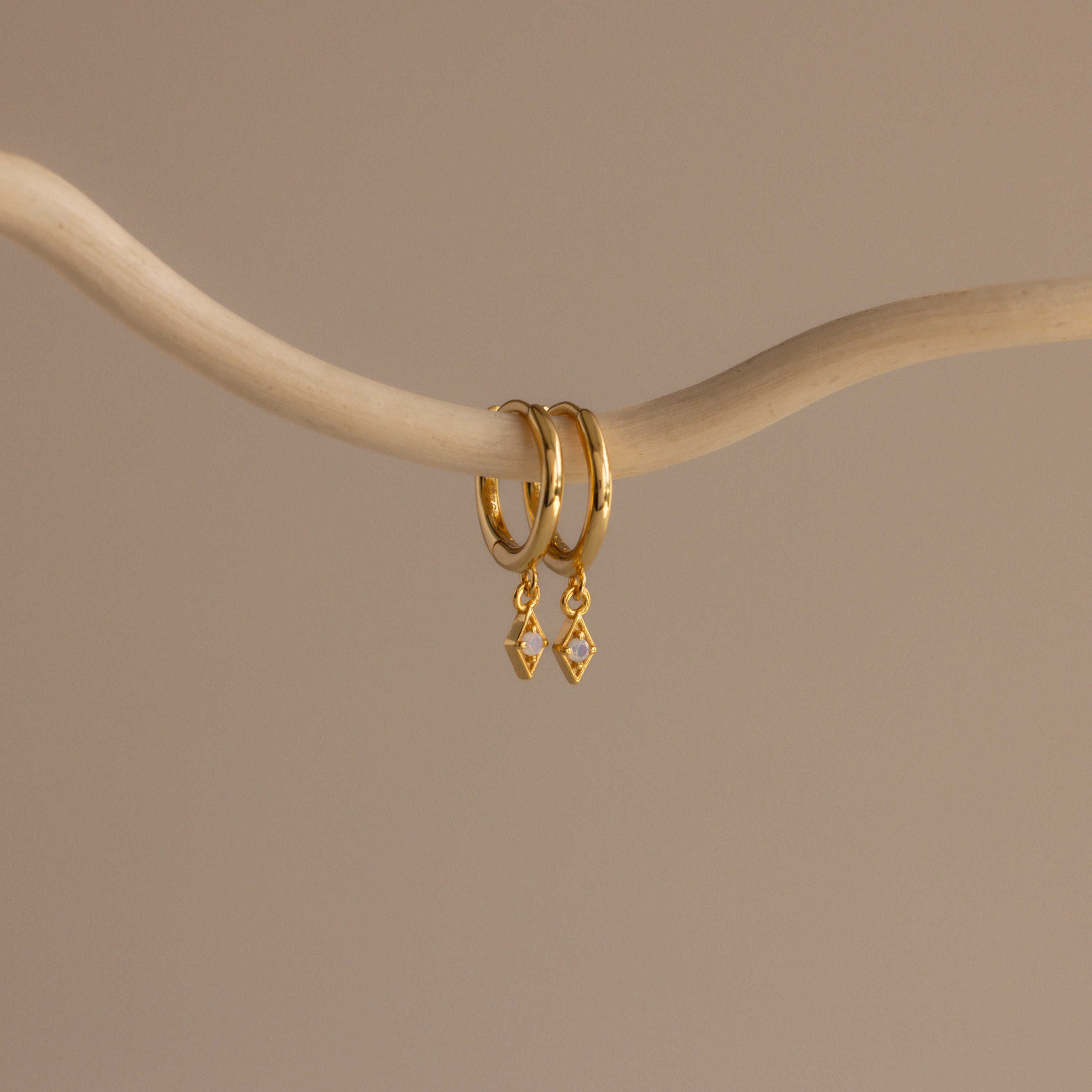 The Ericka Opal Huggies, featuring gold hoops with diamond-shaped pendants, hang on a curved branch—an elegant addition to any personalized jewelry collection.