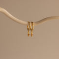 The Ericka Birthstone Huggies in Garnet (January) feature two gold hoops with small diamond-shaped red charms, hanging from a curved beige branch—ideal as birthstone huggies or a unique twist on diamond charm earrings.
