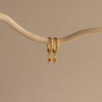 The Ericka Birthstone Huggies in Garnet (January) feature two gold hoops with small diamond-shaped red charms, hanging from a curved beige branch—ideal as birthstone huggies or a unique twist on diamond charm earrings.