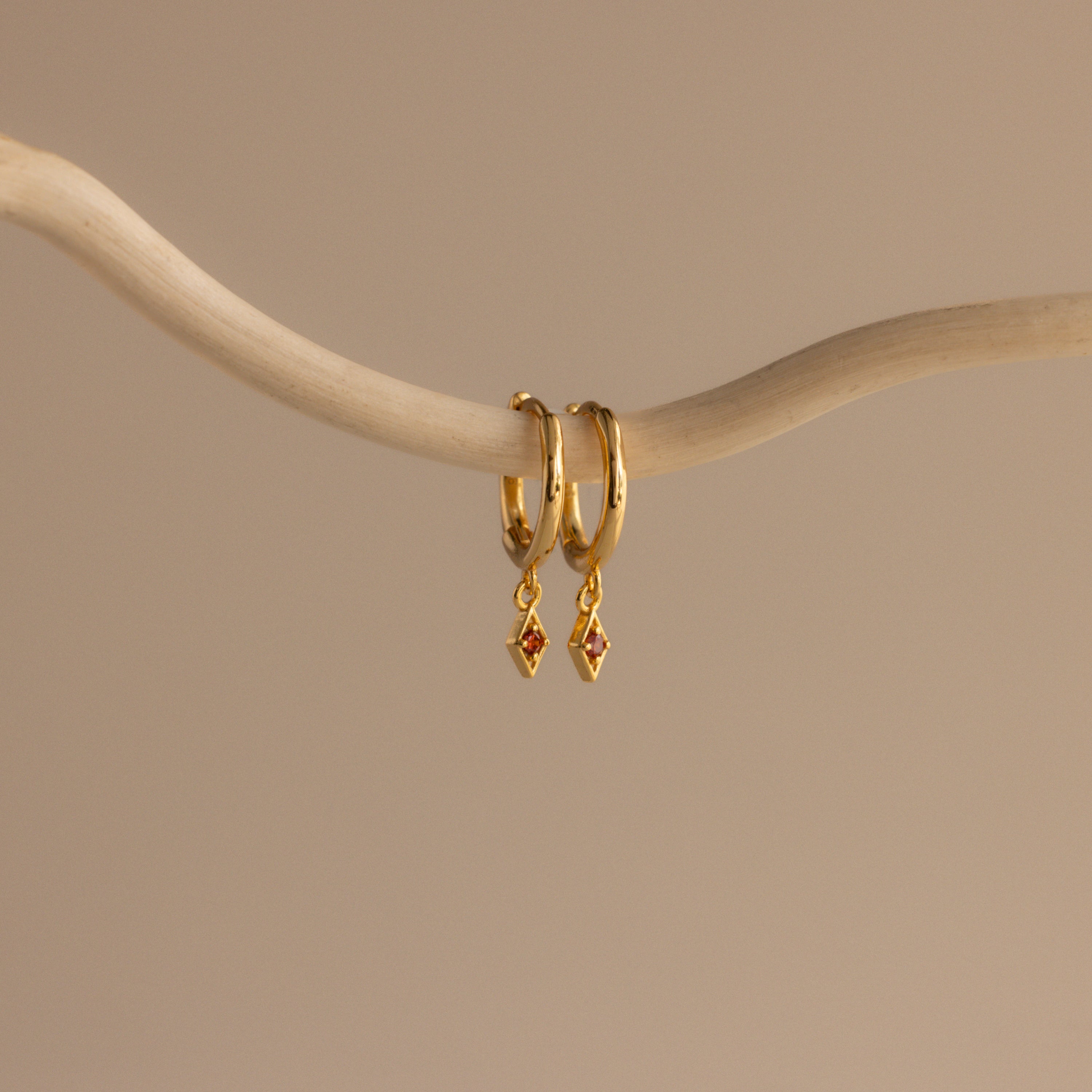 The Ericka Birthstone Huggies in Garnet (January) feature two gold hoops with small diamond-shaped red charms, hanging from a curved beige branch—ideal as birthstone huggies or a unique twist on diamond charm earrings.