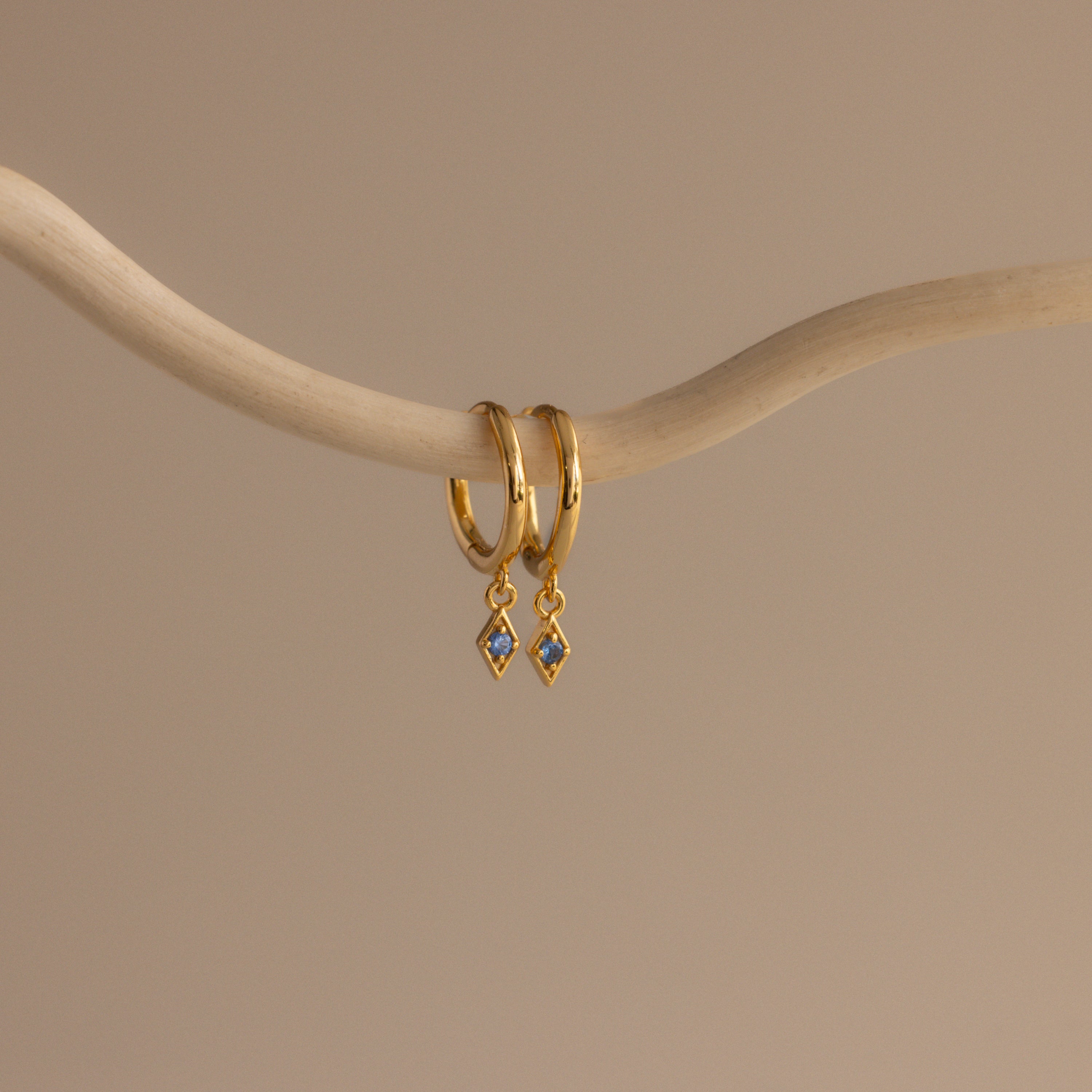 The Ericka Blue Zircon Huggies—gold hoop earrings with blue diamond-shaped zircon pendants—are a stylish way to add sparkle and elegance to your look.