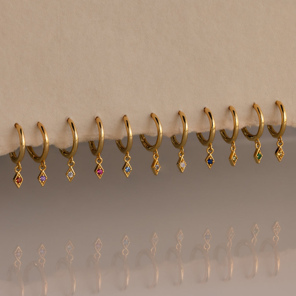 Ten Ericka Birthstone Huggies in Alexandrite (June) rest on a beige surface, their diamond-shaped charms sparkling with colorful reflections—perfect for those who love personalized jewelry.