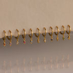 Ten Ericka Birthstone Huggies in Alexandrite (June) rest on a beige surface, their diamond-shaped charms sparkling with colorful reflections—perfect for those who love personalized jewelry.