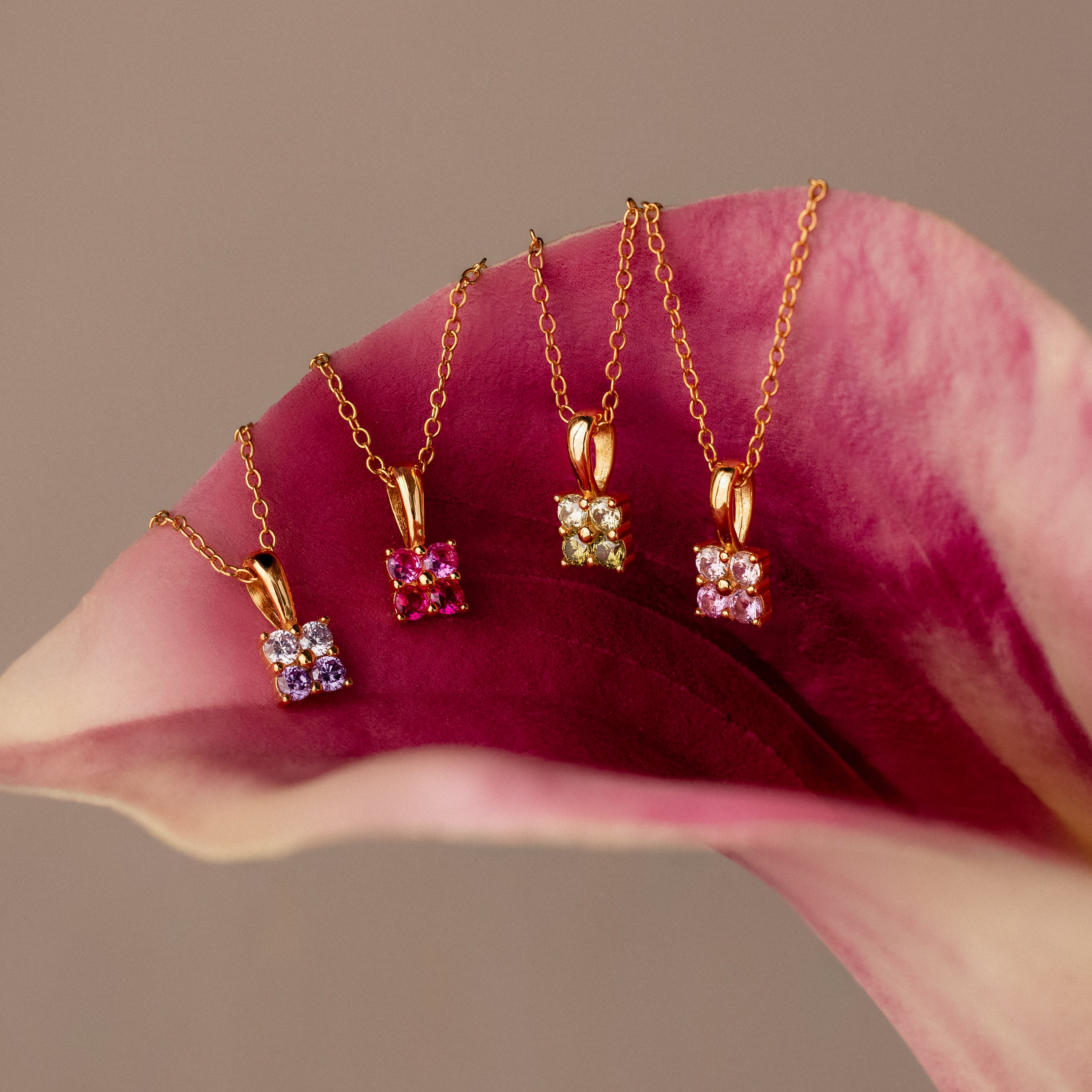 Four gold necklaces with colorful gemstone pendants, including an Ombre Peridot Flower Necklace, displayed on a pink and white calla lily flower.