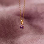 The Ombre Amethyst Flower Necklace features a gold chain with a small flower pendant adorned with purple amethyst gemstones, photographed on a soft pink background.