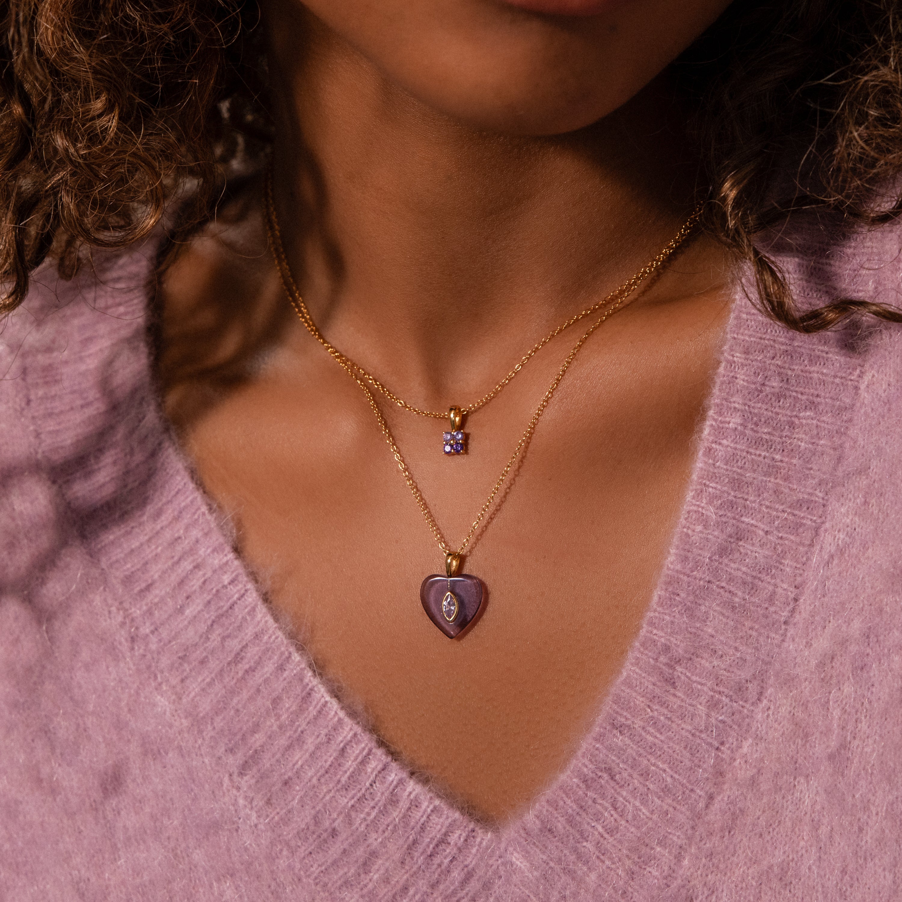 Woman in a lilac sweater wears two gold necklaces: one with a heart-shaped purple pendant and the other featuring our Ombre Ruby Flower Necklace.