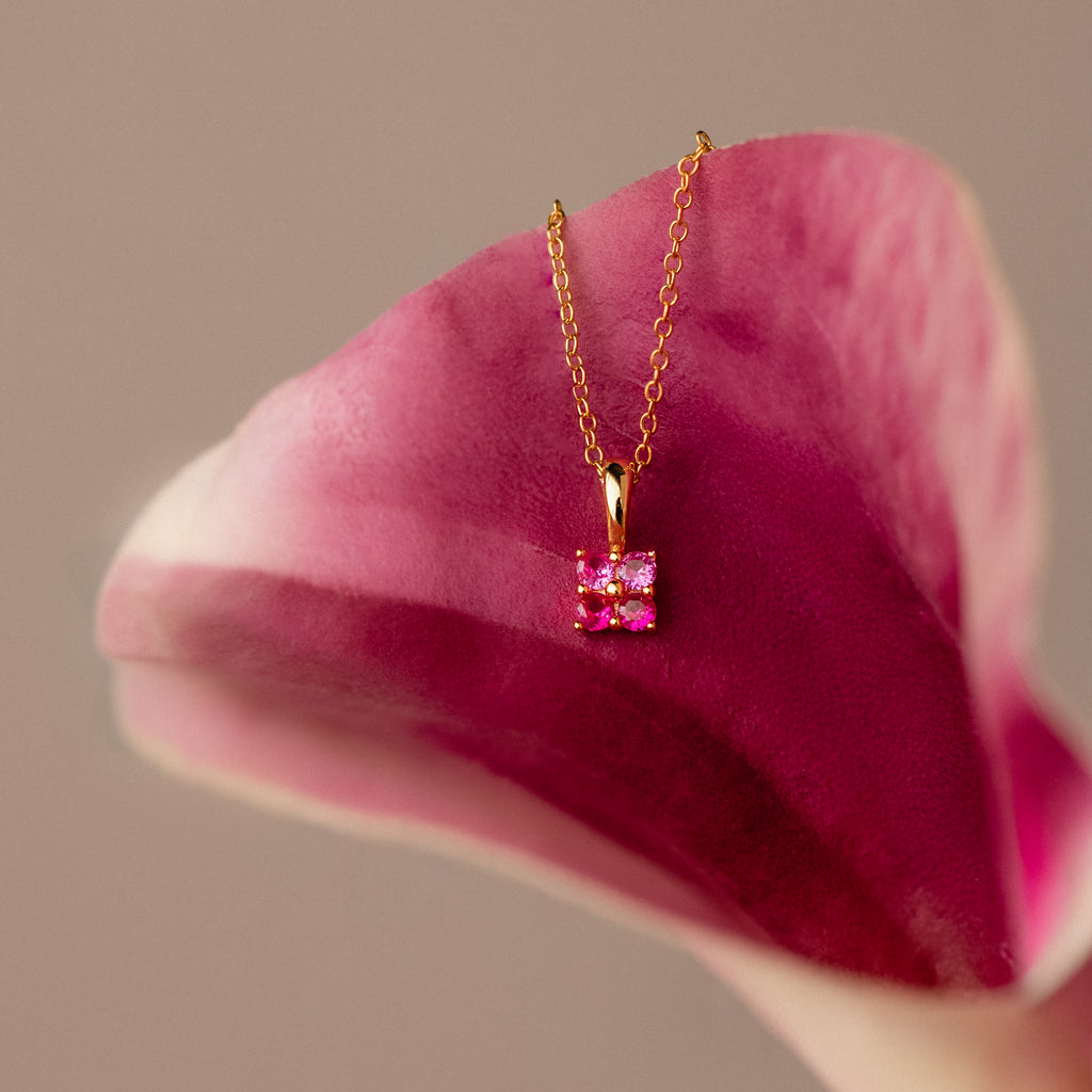 The Ombre Ruby Flower Necklace features a pink gemstone pendant, showcased on pink and white flower petals atop a neutral background.